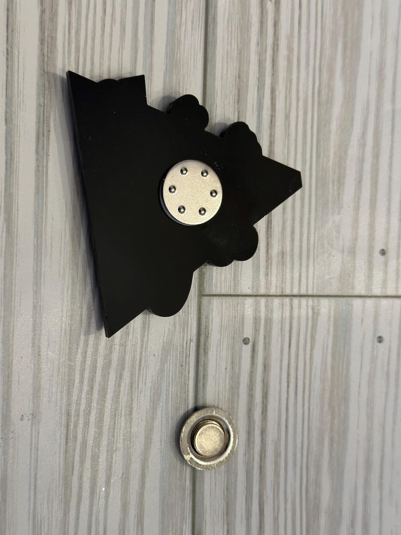 Op de afbeelding: Zwarte en zilveren magnetische pin met een cartoon afbeelding van een huis. De pin heeft een ronde magneet aan de achterkant.