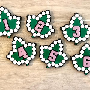May include: Six leaf-shaped ornaments featuring a green center, a white beaded border, and a large pink number in the middle. The numbers one through six are displayed individually on each leaf, with a black backing visible along the edges.