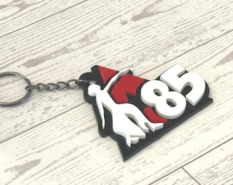 May include: A keychain with a black, red, and white design. The keychain features the number 85 in white, a stylized figure, and a red and black shape. The keychain is attached to a silver keyring.