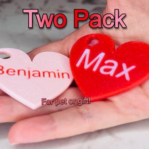 May include: Two heart-shaped pet tags, one pink with the name "Benjamin" and one red with the name "Max". The tags are held in a hand and the text "For pet or gift!" is visible below.
