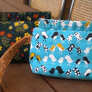 May include: Two fabric bags with different patterns. One bag is dark green with a floral and bee pattern. The other bag is turquoise blue with a white bird pattern. Both bags have brown handles.