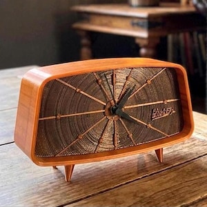 May include: A horizontal, rectangular table clock with a smooth, light brown wooden frame and three small legs. The clock face features a dark, textured, circular pattern with radiating lines and black hands. The word Philco is visible in script.