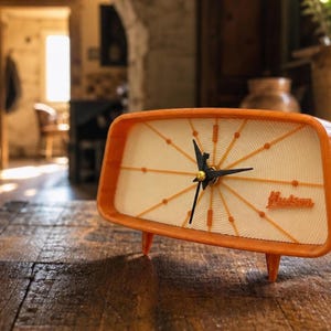 May include: A rectangular orange table clock with a cream-coloured face featuring a sunburst pattern of orange lines and dots. The clock has black hands and the word Hudson printed in orange script on the lower right side of the clock face.
