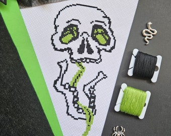 May include: A white cross-stitch banner featuring a skull with green eyes and a green spine. The banner is decorated with black and green thread spools, a silver snake charm, and a spider charm. The banner has a green border.