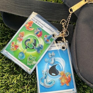 Puede incluir: Dos cartas de intercambio de Pokémon con una cadena dorada unida. La carta de la izquierda es verde con un círculo en el centro y varios personajes de Pokémon alrededor del círculo. La carta de la derecha es azul con un círculo en el centro y tres personajes de Pokémon alrededor del círculo. El texto de la carta de la izquierda dice "Basic Energy".
