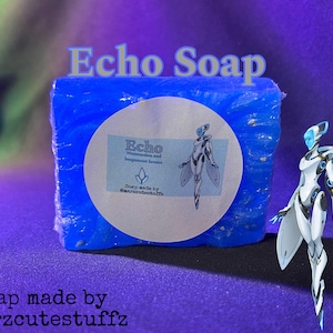 以下が含まれることがあります： 白いラベルが付いた青い石鹸。ラベルには「Echo Watermelon and Bergamot Leaves」と「Soap made by @marzcutestuffz」と書かれており、翼のある女性の漫画が描かれている。