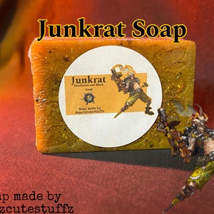 Puede incluir: Una pastilla de jabón de color amarillo y naranja. El jabón está decorado con purpurina y tiene una etiqueta que dice "Junkrat Eucalyptus and Black Sand Soap made by @marzcutestuffz". La etiqueta también presenta un personaje de dibujos animados del videojuego Overwatch.