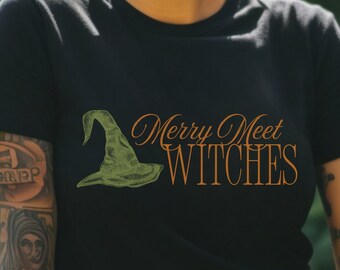 May include: Black t-shirt with the text "Merry Meet Witches" in orange script. The design includes a green witch hat. The person in the image is wearing black glasses and smiling.