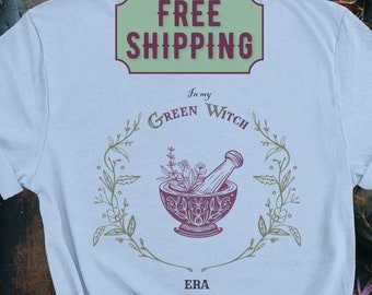 May include: Baby blue t-shirt with a botanical design featuring a mortar and pestle, herbs, and the text "In my Green Witch Era." The design is in shades of purple and green. The shirt is a light blue color.