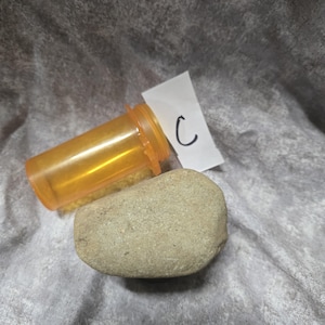 May include: An orange prescription bottle and a smooth, light gray stone are displayed. A small piece of paper with the letter "C" written on it is also present. The items are on a gray, textured surface.