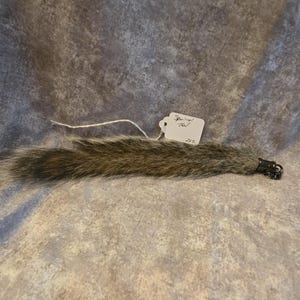 May include: A brown and gray squirrel tail with a dark metal clasp at one end. A white paper tag is attached to the fur with a string, featuring the handwritten text "Squirrel Tail" and the number "222" written in black ink on the tag.