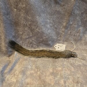 May include: A brown and gray squirrel tail attached to a silver-toned metal cap with a geometric pattern. A small white paper tag is tied to the cap with string, featuring the handwritten text Squirrel Tail and the number 221 in black ink.