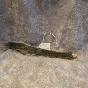 May include: A gray and black squirrel tail with a metallic gold-toned cap at one end. A small white paper tag is attached to the fur, featuring the handwritten text "Squirrel Tail" and the number "228" in black ink, with a thin white string loop.