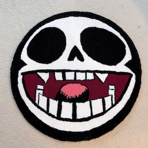 May include: A round rug featuring a cartoon skull design. The rug has a black border, white skull, and black eye sockets. The mouth is open with white teeth, a red interior, and a pink tongue.