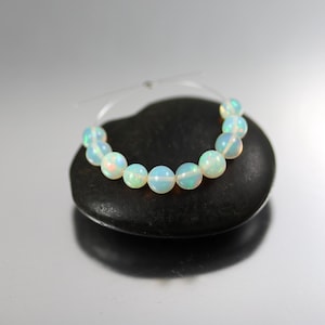 May include: A strand of nine round, translucent white opal beads with iridescent flashes of blue, green, and orange. The beads are strung on a thin, clear filament thread, arranged in a curved line against a smooth, dark, heart-shaped stone.