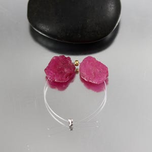 May include: Two raw, deep pink ruby gemstones are strung on a thin, clear nylon cord. A single small, metallic gold-toned spacer bead is positioned between the two stones, and a silver-toned crimp bead secures the ends of the clear nylon wire.