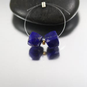 May include: A pair of faceted, deep blue lapis lazuli gemstone beads, strung on a thin wire, with a small gold-colored bead between them. The beads are cube-shaped and reflect light. A silver-colored square is at the top of the wire.