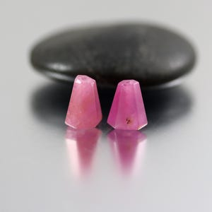 May include: Two small, faceted, trapezoidal-shaped pink gemstone beads. Each bead features a flat top with a visible drill hole and a wider base. The stones have a translucent quality with natural internal inclusions and a polished surface finish.