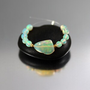 May include: A bracelet featuring a central irregular shaped opal gemstone flanked by small gold beads and a series of round, translucent, iridescent opal beads strung on a thin clear cord, showcasing flashes of green and blue light throughout.