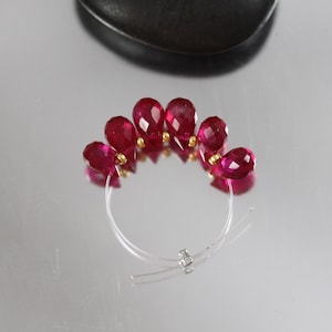 May include: A close-up of a necklace featuring faceted, teardrop-shaped, deep red gemstones. The gemstones are strung on a clear, thin wire, with small gold-colored beads between each stone. The necklace is arranged in a semi-circle.