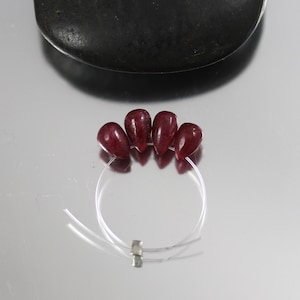 May include: Four deep red, teardrop-shaped beads strung on a clear, thin wire. The beads are arranged in a semi-circle. A small, silver-colored clasp secures the wire. A dark, rounded object is in the background.