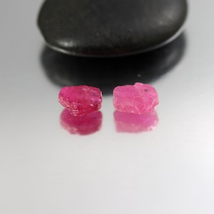 May include: Two rough-cut, vibrant pink ruby gemstones are displayed, reflecting on a shiny surface. A dark, smooth stone sits in the background. The rubies have a raw, natural appearance, showcasing their crystalline structure and rich color.