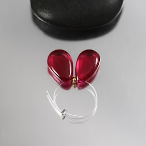 May include: A pair of teardrop-shaped, translucent red earrings with a gold-colored connector. The earrings are strung on a thin, clear wire. A dark, rounded object is in the background.