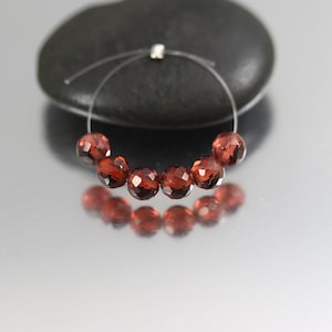 May include: A strand of faceted, deep red garnet beads strung on a thin, clear wire. The beads are arranged in a curved line, with a dark, smooth stone in the background. The beads reflect light, creating a sparkling effect.