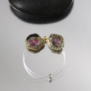 May include: A necklace featuring two raw, semi-precious tourmaline stones with pink centers, black edges, and beige exteriors. The stones are strung on a clear, thin cord with a gold bead and a silver clasp. The necklace is displayed on a reflective surface.