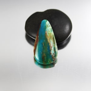 May include: A teardrop-shaped gemstone with a polished surface. The stone features a blend of turquoise, teal, and brown hues, with streaks and patterns. A dark, rounded stone sits behind the gemstone, providing contrast.