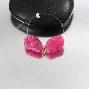 May include: A pair of raw, hot pink ruby gemstone earrings. The earrings are strung on a thin, clear wire with a small silver clasp. Each earring features a rough-cut ruby with a gold bead accent.