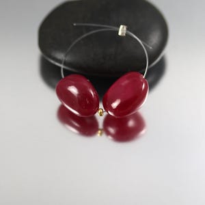 May include: A pair of red, oval-shaped earrings with a wire and gold-colored accents. The earrings are displayed on a reflective surface with a dark, rounded stone in the background. The earrings have a polished, glossy finish.