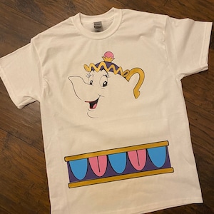 May include: White t-shirt with a cartoon image of Mrs. Potts, the teapot from Disney's Beauty and the Beast. The image shows Mrs. Potts' face with a pink and purple crown and a gold teapot spout. The bottom of the shirt features a gold band with purple, pink, and blue accents.