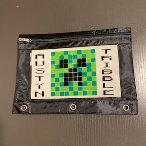 May include: Black zippered pencil case with a clear plastic window. The window shows a green and black pixelated Minecraft Creeper face and the names "Austin" and "Tribble" written in black.