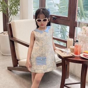 May include: A young girl wearing a light blue sleeveless dress with a white collar and a small blue pocket. The dress has a white and blue floral pattern. She is wearing white socks and black shoes. She is also wearing white oval sunglasses.