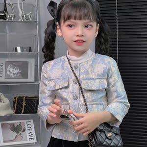 Può includere: Una bambina che indossa una giacca blu chiaro con un motivo floreale e una gonna nera. Tiene in mano una piccola borsa nera con una tracolla a catena dorata. La giacca ha una chiusura a bottone e una tasca sul davanti. La bambina ha i capelli neri con un fiocco nero.