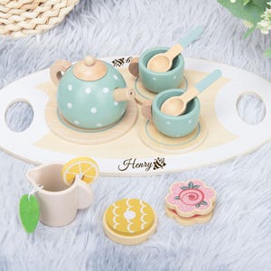 Puede incluir: Un juego de té de juguete de madera con una tetera y tazas de color azul claro con lunares blancos. El juego incluye una bandeja, cucharas, una crema con una rodaja de limón y dos pasteles decorativos. La marca "Henry" está impresa en la bandeja.