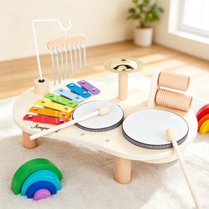 Könnte beinhalten: Ein musikalischer Aktivitätstisch aus Holz mit einem bunten Xylophon, zwei weißen Trommeln, einem goldfarbenen Becken, einem hängenden Glockenspiel und zwei Holzschlägeln. Die Tischoberfläche ist aus hellem Holz mit dem Markennamen Jasper.