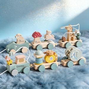Puede incluir: Un juego de tren de juguete de madera con varios vagones conectados en colores pastel y acabados de madera natural. Los vagones incluyen figuras de un oso, un hongo, un conejo, una jirafa, una estrella y una ballena, con los nombres Emma y Olivia.