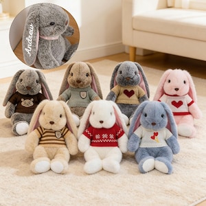 May include: A collection of seven plush stuffed rabbits with long ears, each wearing a knitted jumper. The rabbits are in various colours including grey, beige, white, and pink. One grey rabbit features the name Andrew embroidered on its ear.