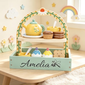 Può includere: Un set da tè giocattolo in legno a due piani con una teiera gialla decorata con margherite, una tazza gialla, un cupcake marrone, una fetta di torta marrone, due dolcetti colorati e un biscotto. La base verde chiaro riporta il nome Amelia.