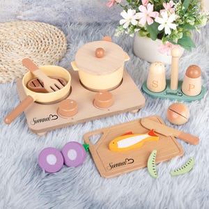 Puede incluir: Juego de cocina de juguete de madera con acabado en madera clara. Incluye una sartén, una olla, una tabla de cortar con un pez, saleros y pimenteros y verduras. El nombre "Sienna" está impreso en varias piezas.