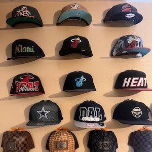 Puede incluir: Una colección de gorras de béisbol expuestas en una pared. Las gorras presentan varios diseños, logotipos y combinaciones de colores, incluidos logotipos de equipos y telas estampadas. Las gorras son de varios colores, incluyendo negro, marrón y azul.