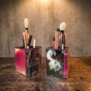 May include: Two square pencil holders made from repurposed cassette tapes, each featuring album art from "The Cure." One is pink and red, the other green and white. Both hold brushes and pencils, perfect for a desk or vanity.
