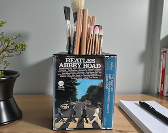 May include: A rectangular desk organizer constructed from clear plastic cassette cases featuring the Beatles Abbey Road album cover art on the front and Sgt. Pepper's Lonely Hearts Club Band on the side, filled with various makeup and art brushes.