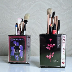 May include: Two makeup brush holders crafted from recycled cassette tapes. One showcases the "Songs of Faith and Devotion" album art by Depeche Mode. The other features the "Violator" album art. Both contain makeup brushes and pencils.