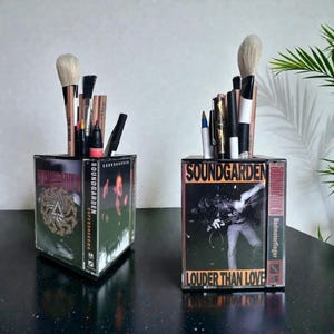 May include: Two Soundgarden cassette tape holders, each filled with makeup brushes and pens. The holders feature album art and text, including "Louder Than Love" and "Superunknown". The holders are on a dark surface.
