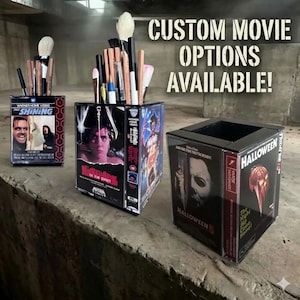 May include: Three black and clear boxes made from VHS tapes, each holding makeup brushes. The boxes feature movie titles: "The Shining", "Nightmare on Elm Street", and "Halloween 5". The text "CUSTOM MOVIE OPTIONS AVAILABLE!" is visible.