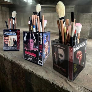 May include: Three square desk organisers made from vintage VHS cases featuring horror movie covers for The Shining, A Nightmare on Elm Street, Halloween 5, and Halloween. Each container is filled with various makeup brushes and writing pens.
