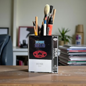 May include: A black and white cassette tape repurposed as a pen holder, featuring the band Rush's album "2112" and "A Farewell to Kings." The holder contains various art supplies, including pencils, brushes, and markers. The cassette tape is on a wooden surface.